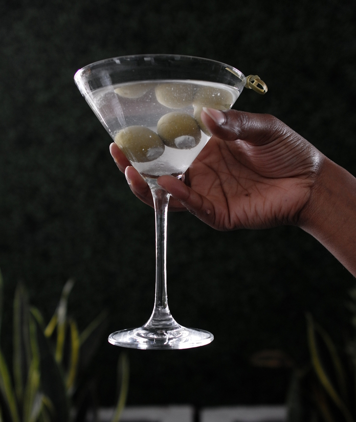 Hand holding a martini glass with olives, dark background, blurred greenery.
