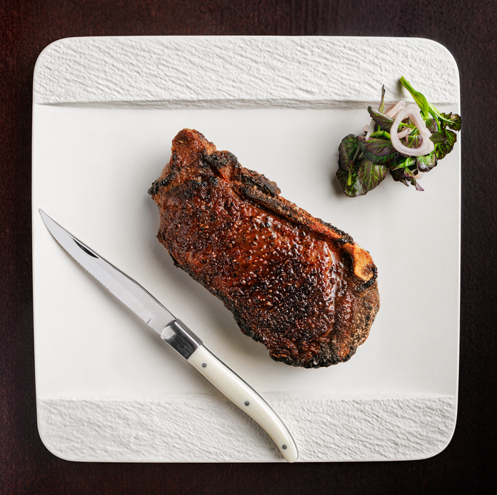 Grilled bone-in steak with charred crust on white square plate, accompanied by sautéed greens and a steak knife