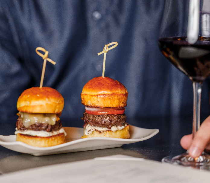 Two gourmet beef sliders with knotted bamboo skewers on a white plate, served alongside a glass of red wine.